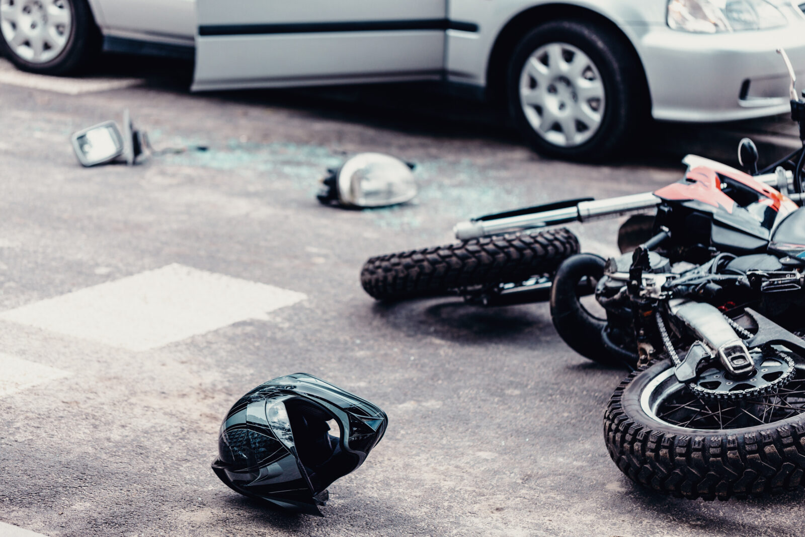 Motorcycle lying on the roadway after a collision with a nearby car, with helmets, broken glass, and debris scattered across the accident scene.